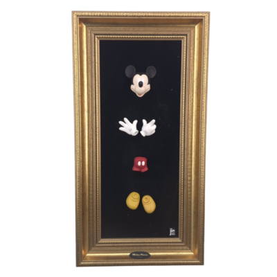 Vertical gold-framed display on black background showing detachable Mickey Mouse parts: black ears, white gloves, yellow shoes, and red shorts with white buttons.